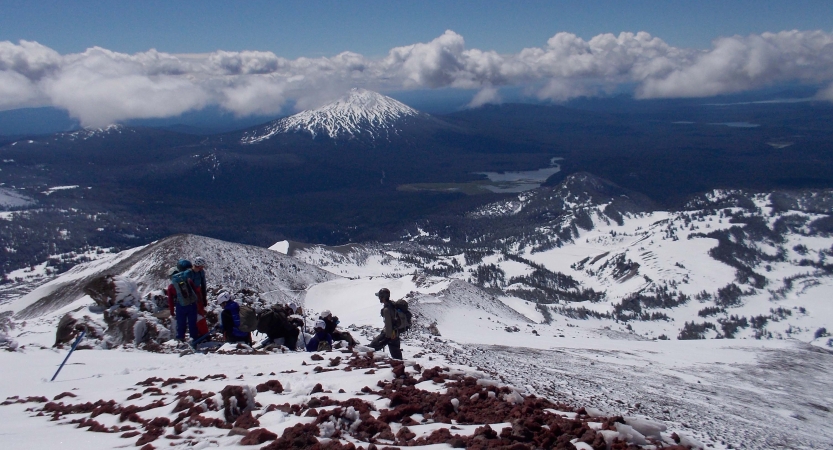 A person wearing a backpack stands on a snowy and rocky outlook, looking out over a vast mountainous landscape.